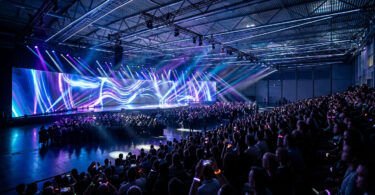 Salle de conférence plongée dans l'obscurité avec un écran géant affichant des lumières néon bleues et violettes devant la foule.