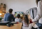 Une famille joue sur une console Switch tandis qu'une personne tient une PS5 en cadeau en arrière-plan.