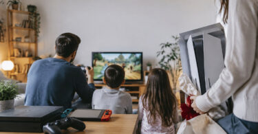 Une famille joue sur une console Switch tandis qu'une personne tient une PS5 en cadeau en arrière-plan.
