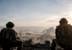 Deux soldats en équipement militaire observant une ville dévastée et enfumée depuis une hauteur.
