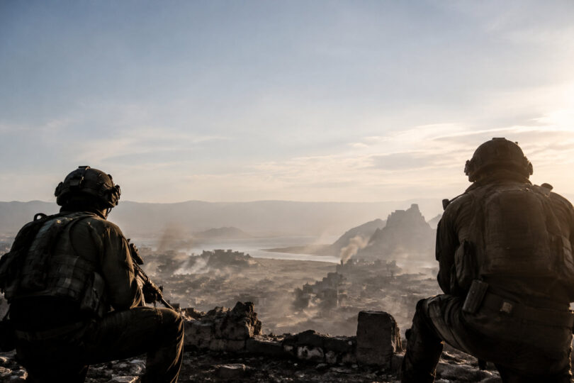 Deux soldats en équipement militaire observant une ville dévastée et enfumée depuis une hauteur.